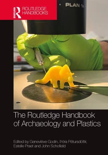 A 3D-printed yellow plastic dinosaur on a table is being touched by a person wearing green gloves. The book cover reads, "The Routledge Handbook of Archaeology and Plastics.